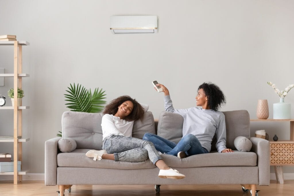 Une mère règle la température de la thermopompe murale, à l'aide de la télécommande, assise sur le canapé avec sa fille.