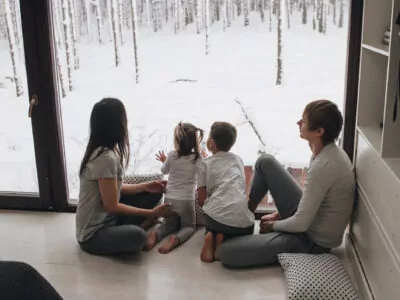 Jeune famille assise à l'intérieur de la maison en hiver, devant un grand mur fenêtre donnant sur une forêt enneigée.