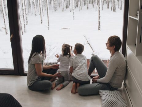 Jeune famille assise à l'intérieur de la maison en hiver, devant un grand mur fenêtre donnant sur une forêt enneigée.
