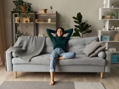 Jeune femme souriante, pieds nus, confortablement assise sur un canapé du salon, les bras dans les airs, une jambe repliée