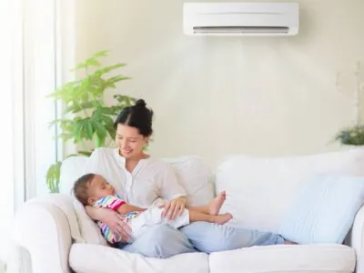 Une mère souriante assise sur un canapé tient son bébé endormi, profitant de la fraicheur du climatiseur