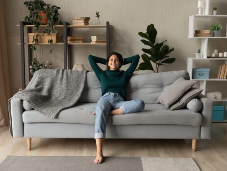 Smiling young woman, barefoot, comfortably seated on a sofa in the living room, arms in the air, one leg bent.